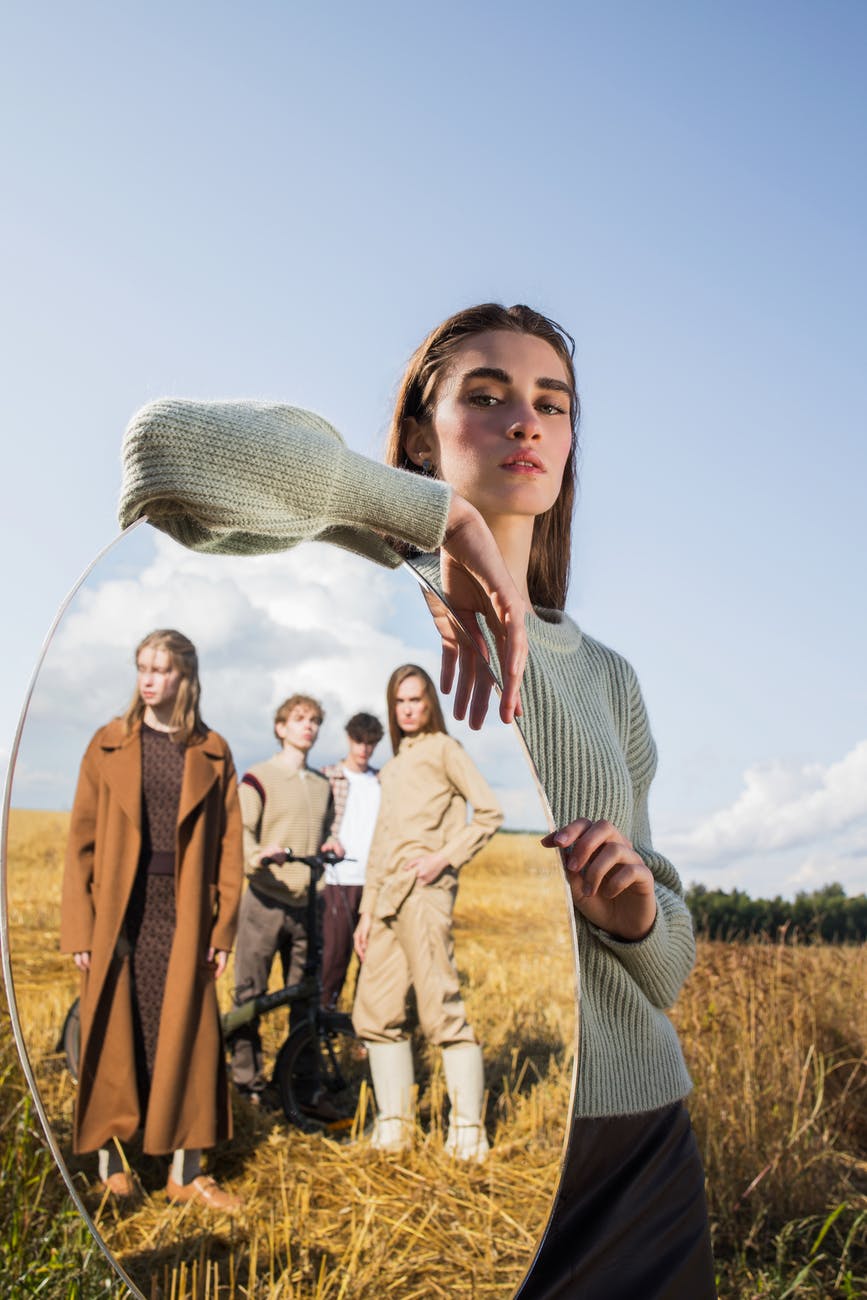young woman standing in field with oval mirror reflecting group of young people