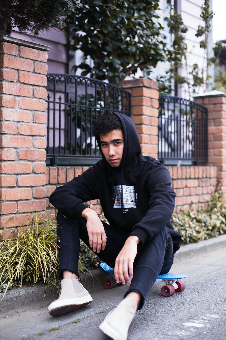 young man wearing black hood sitting on a skateboard by a brick fence