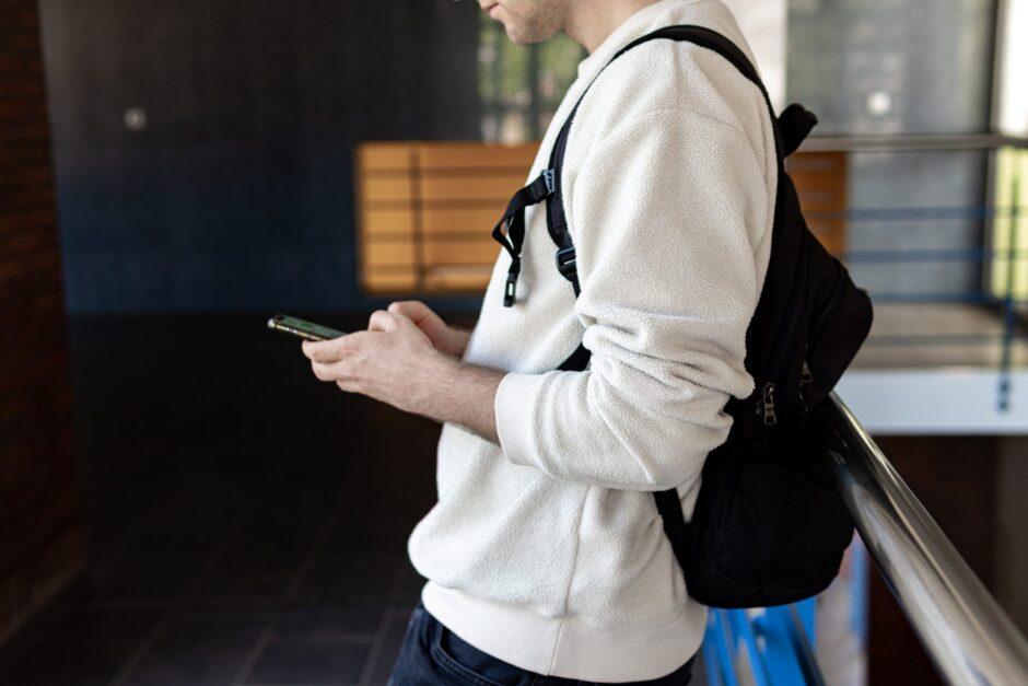 A man in a white shirt and black backpack leans against a chrome railing inside a building while looking at his phone. The man appears relaxed, with his weight resting on one leg and his other foot crossed over it. He holds the phone in one hand and seems to be scrolling through its contents.