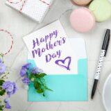 happy mothers day card beside pen macaroons flowers and box near coffee cup with saucer